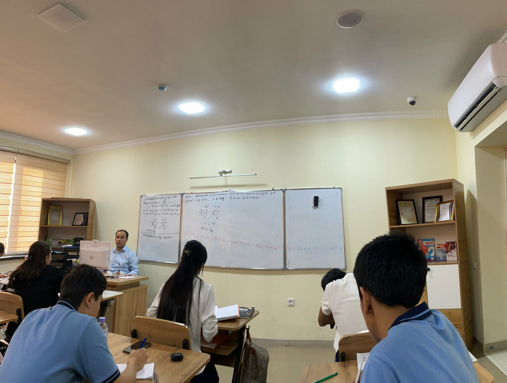 A math lesson: students in light blue polos, a teacher at the board, equations on whiteboards