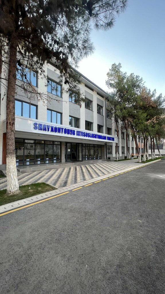 Front of Shayxontohur specialized school: a four-story white and grey building, blue sign, pines, and a striped walkway